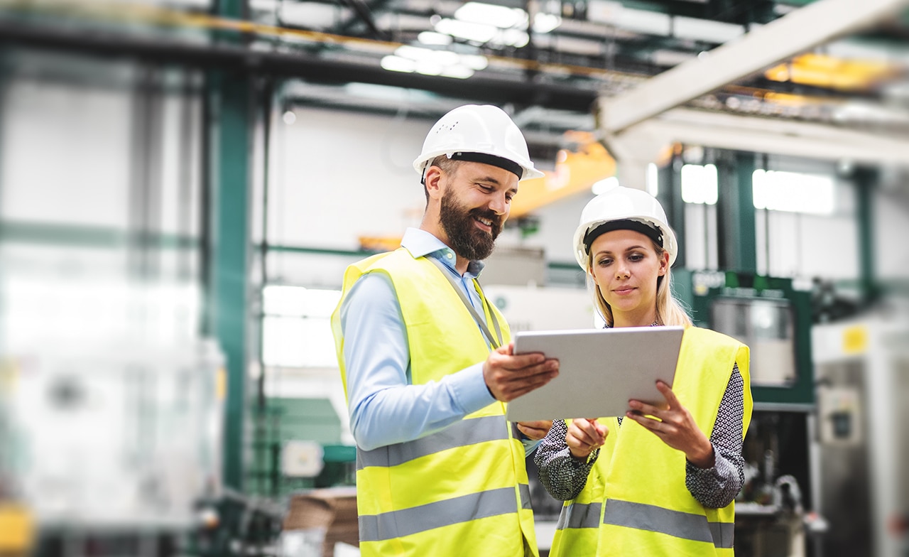 Two people wearing safety vests and safety helmets looking at tablet inside a factory