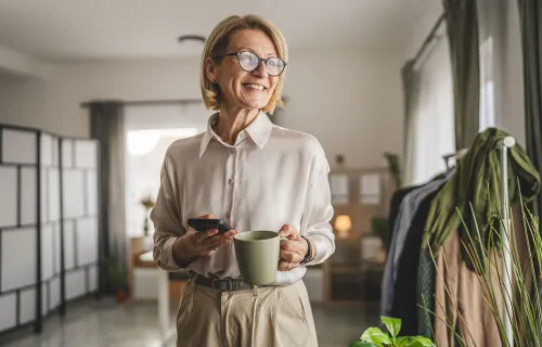 Äldre kvinna med glasögon står inomhus, håller en kopp kaffe och en mobiltelefon, och ler.
