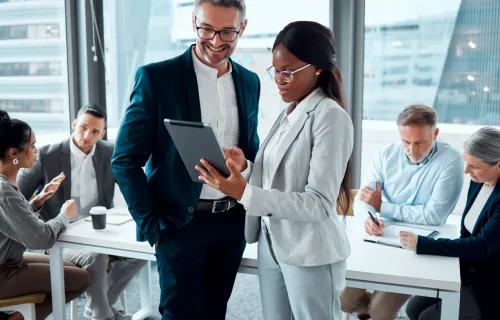 Two colleagues talking while looking at a tablet in a crowded meeting room